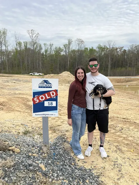 Katie and David holding their small black-and-tan dog beside a SOLD sign on a cleared homesite