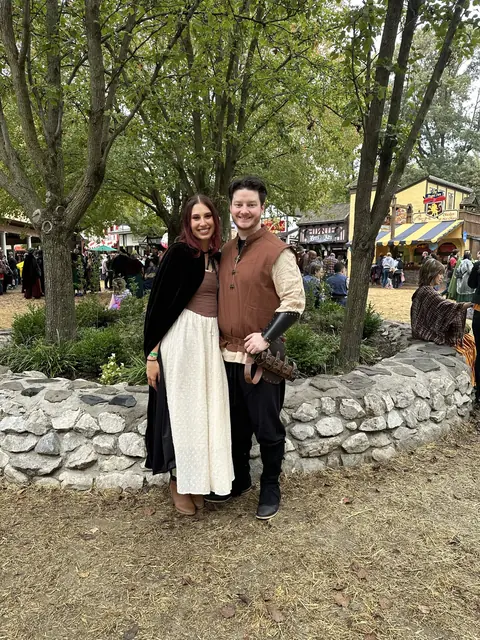 Katie and David in autumn renaissance costumes in front of a low stone wall and tall trees
