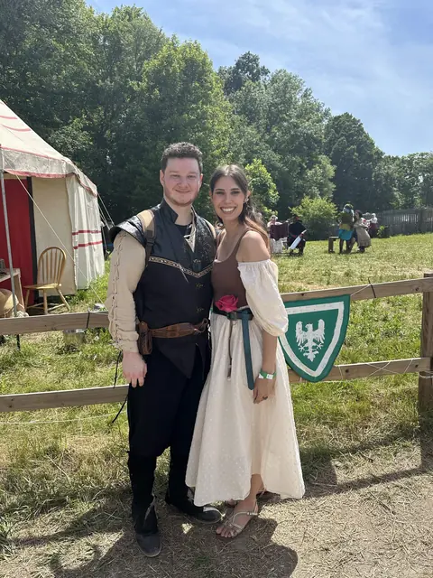 Katie and David in renaissance faire costume beside a wooden fence and a green heraldic banner