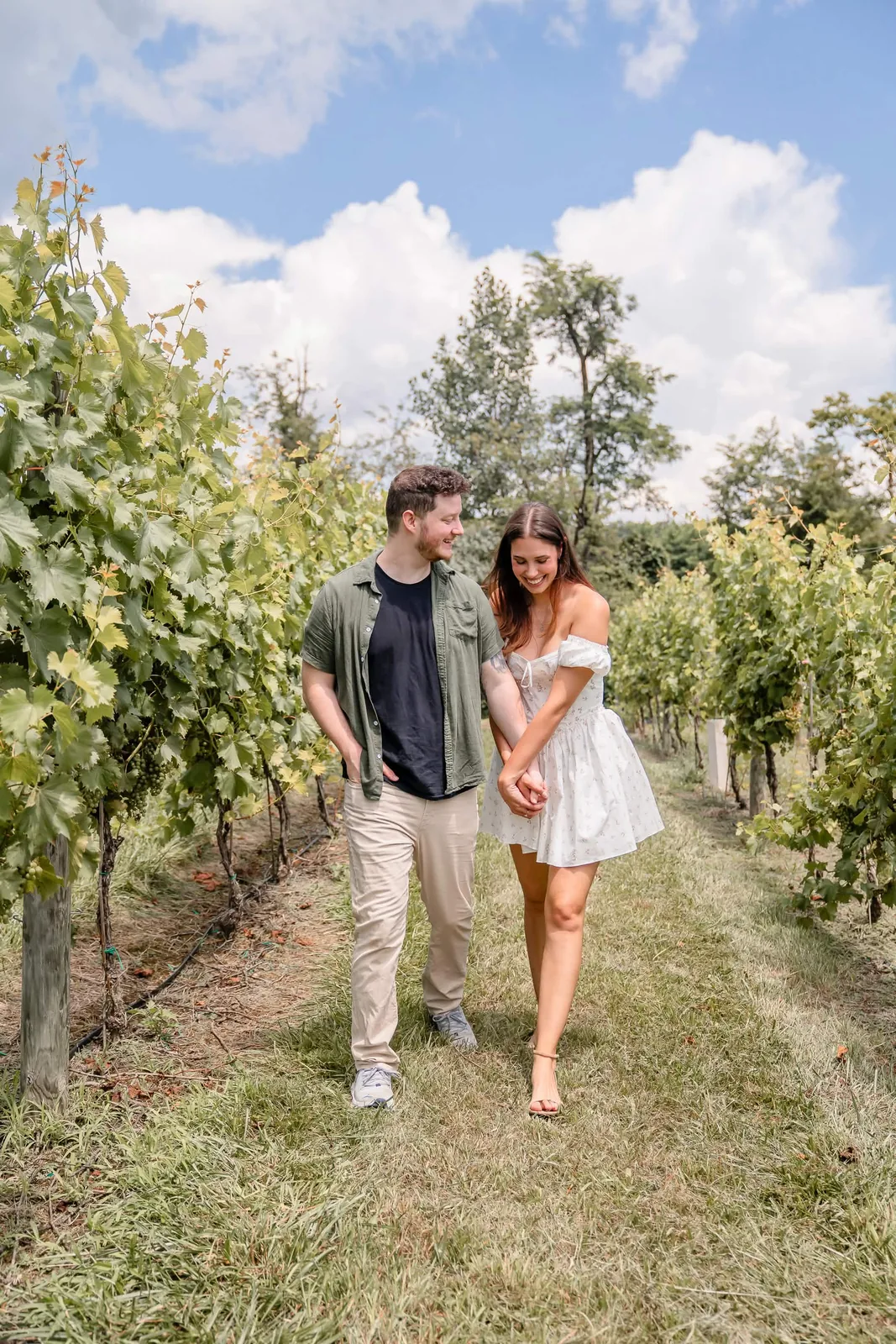 Katie and David walking hand in hand between rows of grapevines under a blue sky