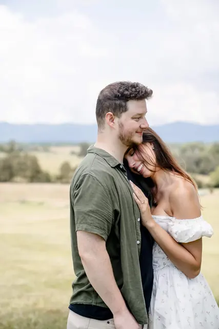 Katie resting her head on David's chest in an open field, mountains behind them, her ring hand on his shirt